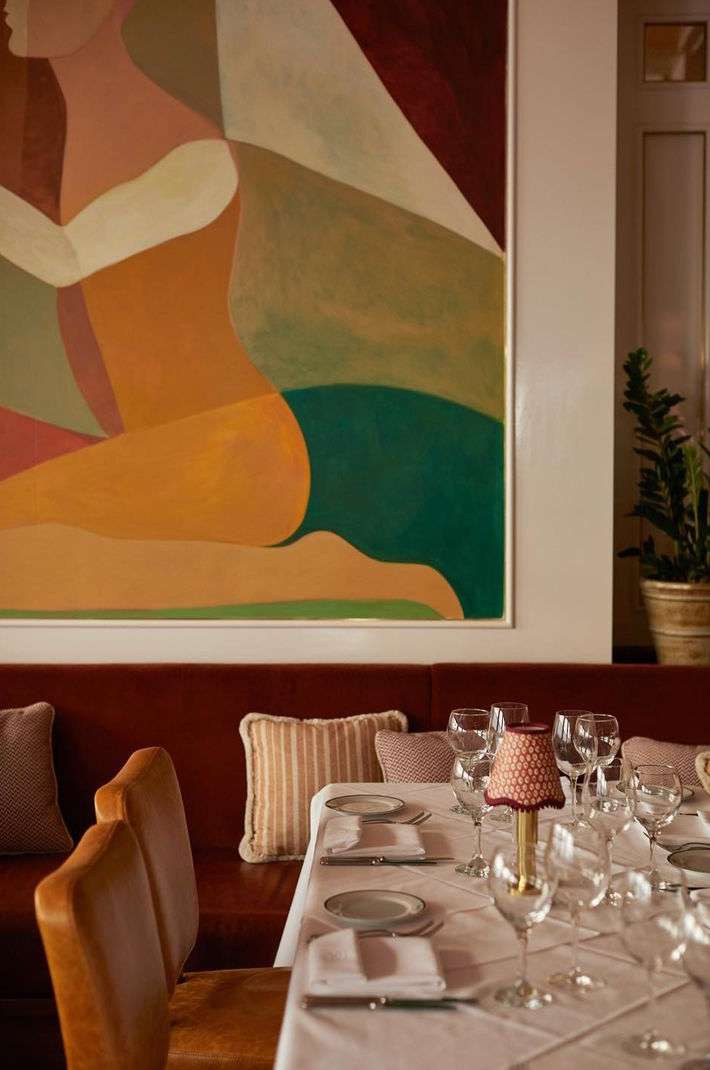 Interior of a restaurant with a set table and abstract art on the wall, featuring a red banquette and upholstered chairs in a warm, inviting space.