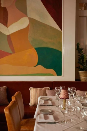 Interior of a restaurant with a set table and abstract art on the wall, featuring a red banquette and upholstered chairs in a warm, inviting space.