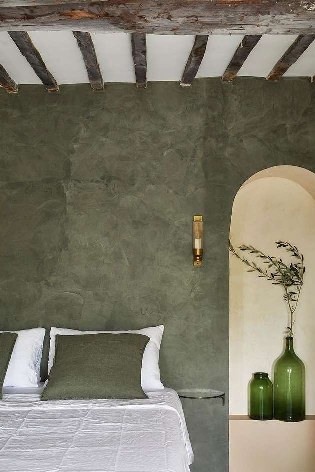 Bedroom interior with a bed, green walls, wooden beams, arched doorway, vases, and olive branches providing a calming aesthetic.