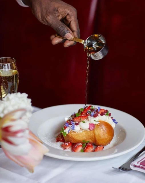 Dark-skinned hand pours liquid over cream-topped baba au rhum dessert garnished with sliced strawberries and flowers, sitting on a white plate.