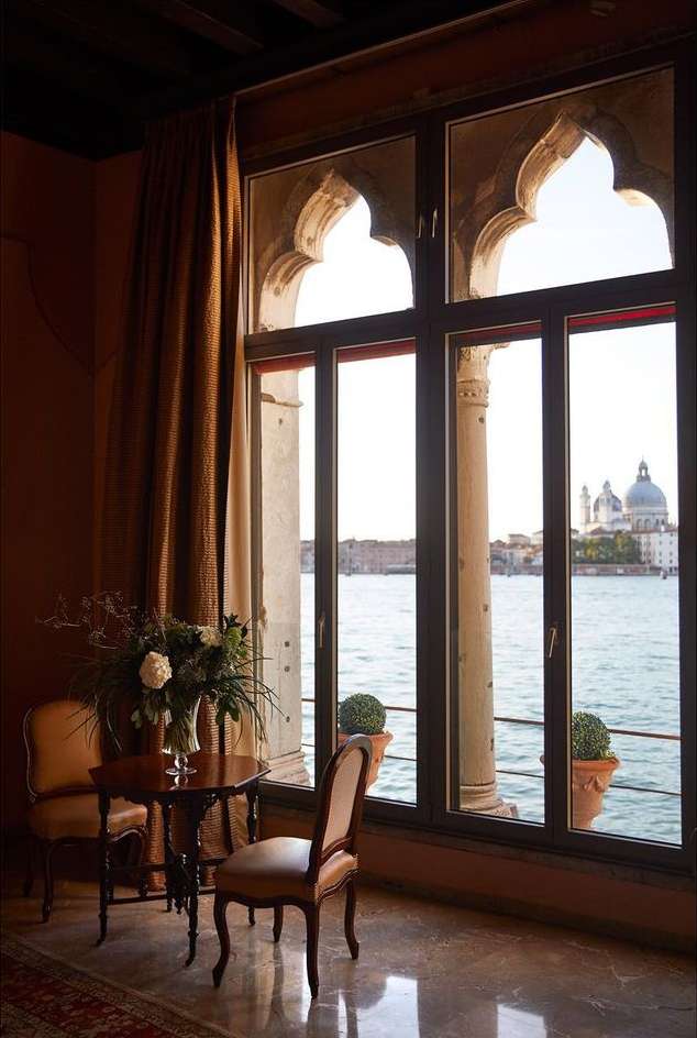 Ornate window frames a serene Venice canal view with water, architecture, and two empty chairs near a small flower-filled table.