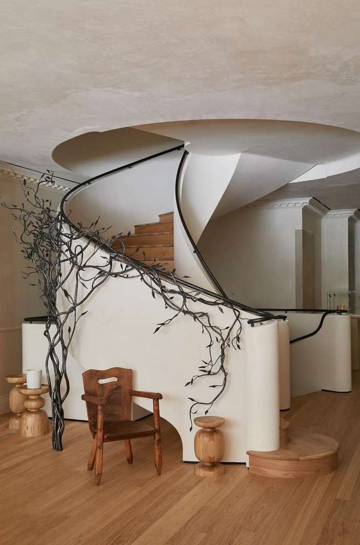 Indoor view of an ornate spiral staircase, wooden chair, and side tables. The modern design features cream walls and hardwood floors.