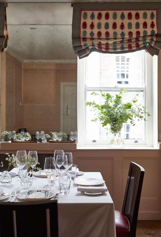 Elegant table setting with wine glasses and white tablecloth, complemented by a window with plants and muted wall tones.