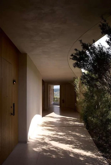Indoor architectural shot of a long hallway with light stone floors, neutral walls and ceiling, wood door, and greenery at the end.