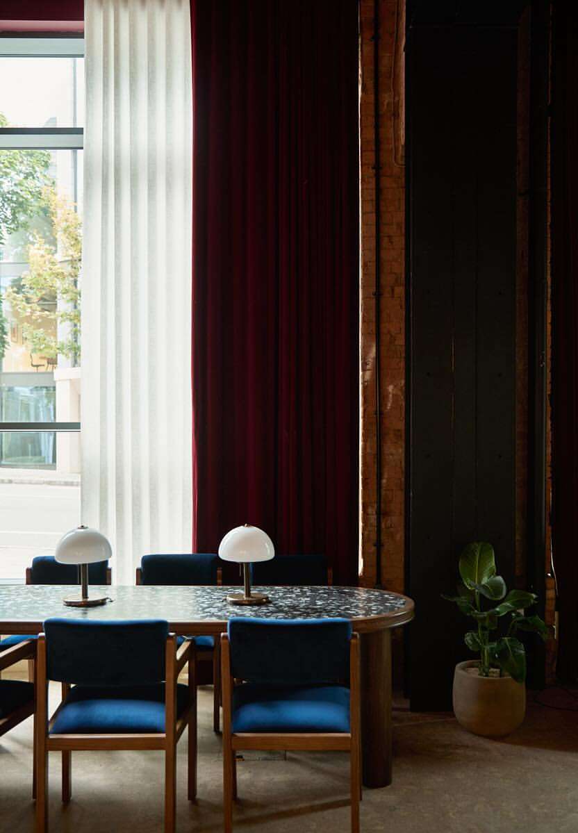 Interior shot of a long dining table with blue chairs and table lamps against tall windows with rich curtains.