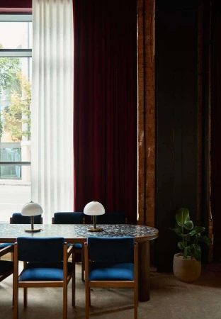 Interior shot of a long dining table with blue chairs and table lamps against tall windows with rich curtains.