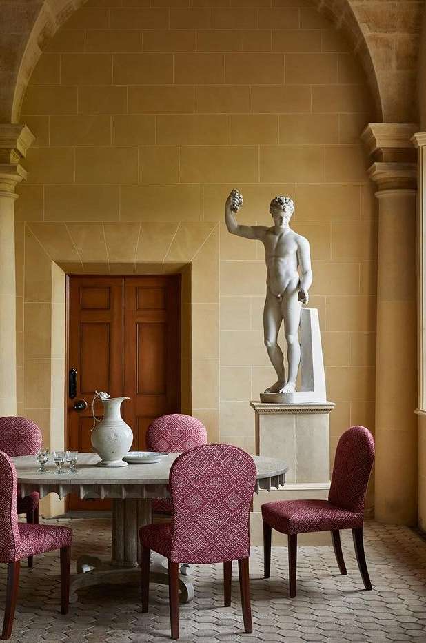 Interior architectural shot of a statue and dining set. Stone walls and floors create a neutral backdrop for red patterned chairs.