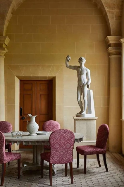 Interior architectural shot of a statue and dining set. Stone walls and floors create a neutral backdrop for red patterned chairs.