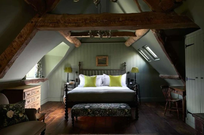 Charming bedroom interior features a wooden four-poster bed with light green pillows, dresser, and wooden beams in an attic setting.