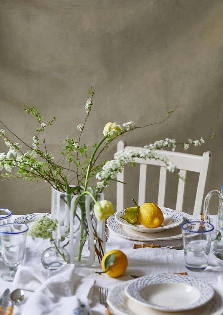 Still life of a table set with white plates, glassware with blue rims, lemons, and white flowers in vases against a neutral backdrop.