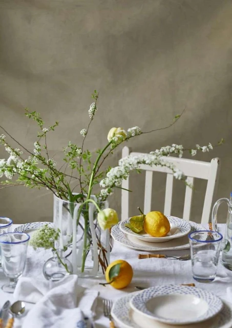 Still life of a table set with white plates, glassware with blue rims, lemons, and white flowers in vases against a neutral backdrop.