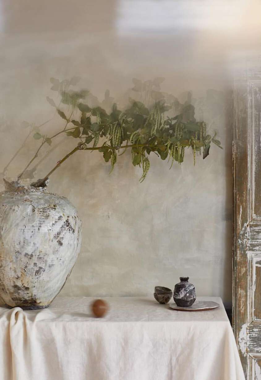 Still life featuring a white linen-covered table, a textured vase with foliage, ceramic vessels, a plate, and an unidentified round object.