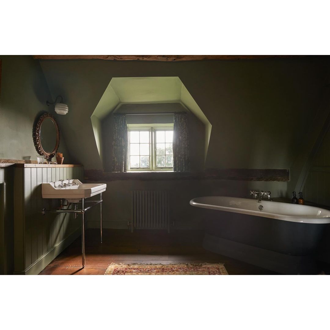 Interior of an olive green bathroom featuring a classic bathtub, sink, oval mirror, window, radiator, and hardwood floors.