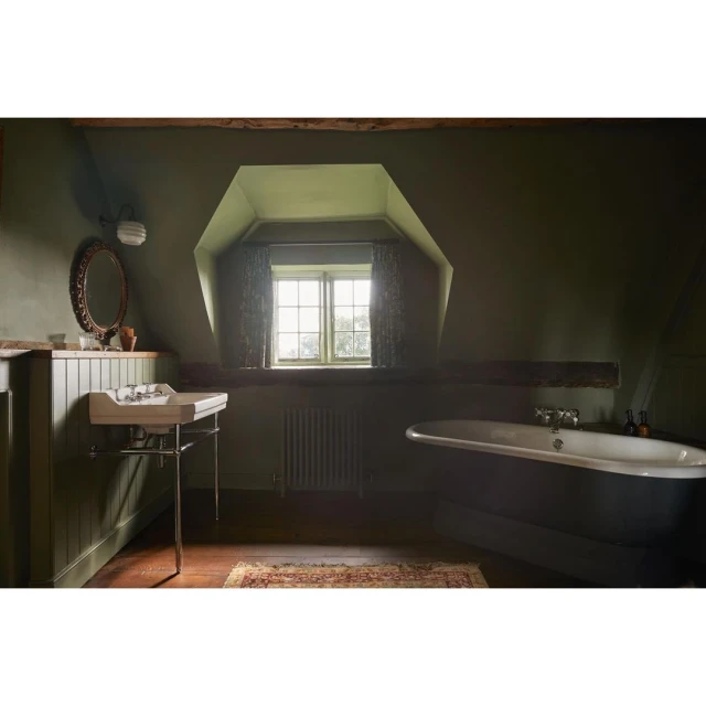 Interior of an olive green bathroom featuring a classic bathtub, sink, oval mirror, window, radiator, and hardwood floors.