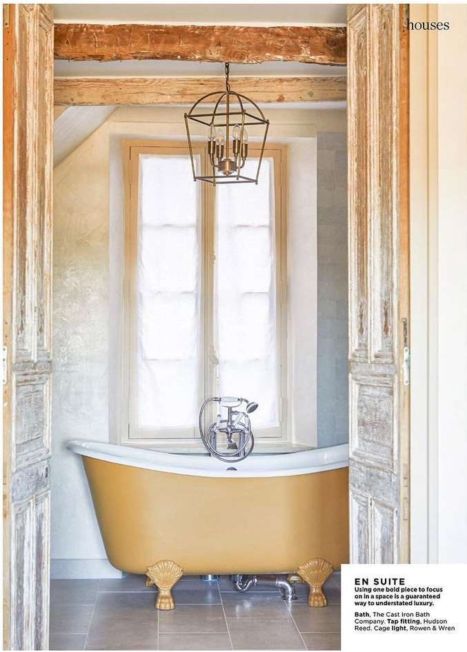 Freestanding gold-colored bathtub in a brightly lit bathroom featuring a window, framed by weathered white wooden doors. 