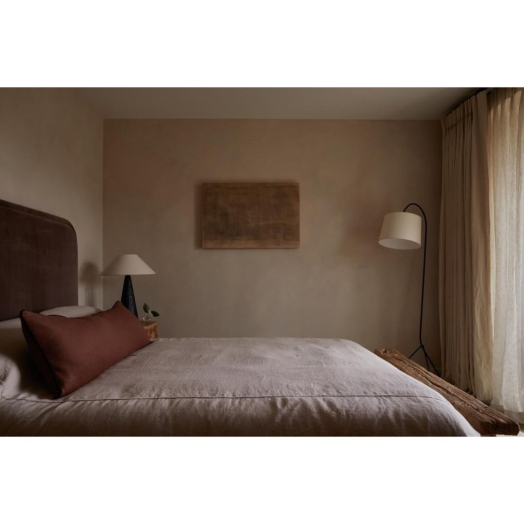 Minimalist bedroom with a queen bed, linen bedding, and a dark headboard. An abstract painting hangs above the bed in a neutral-toned room.