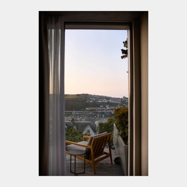 View from a light-filled room onto a waterfront town. A chair and table sit on a patio overlooking the town, boats and water.