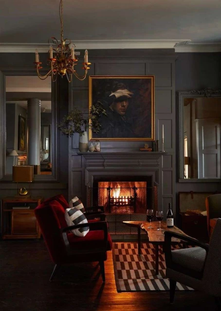 Interior of a room with a fireplace, portrait art above mantel, red velvet chairs, patterned rug, and chandelier lighting overhead.
