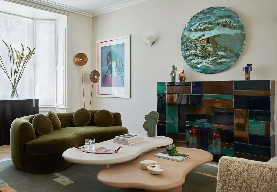 Living room interior with a green velvet sofa, unique coffee tables, and colorful tiled fireplace with a round decorative wall hanging.
