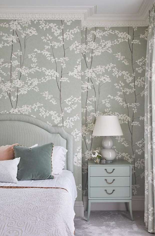 Elegant bedroom interior with a light blue bedside table, a white lamp, and wallpaper featuring a delicate floral pattern.