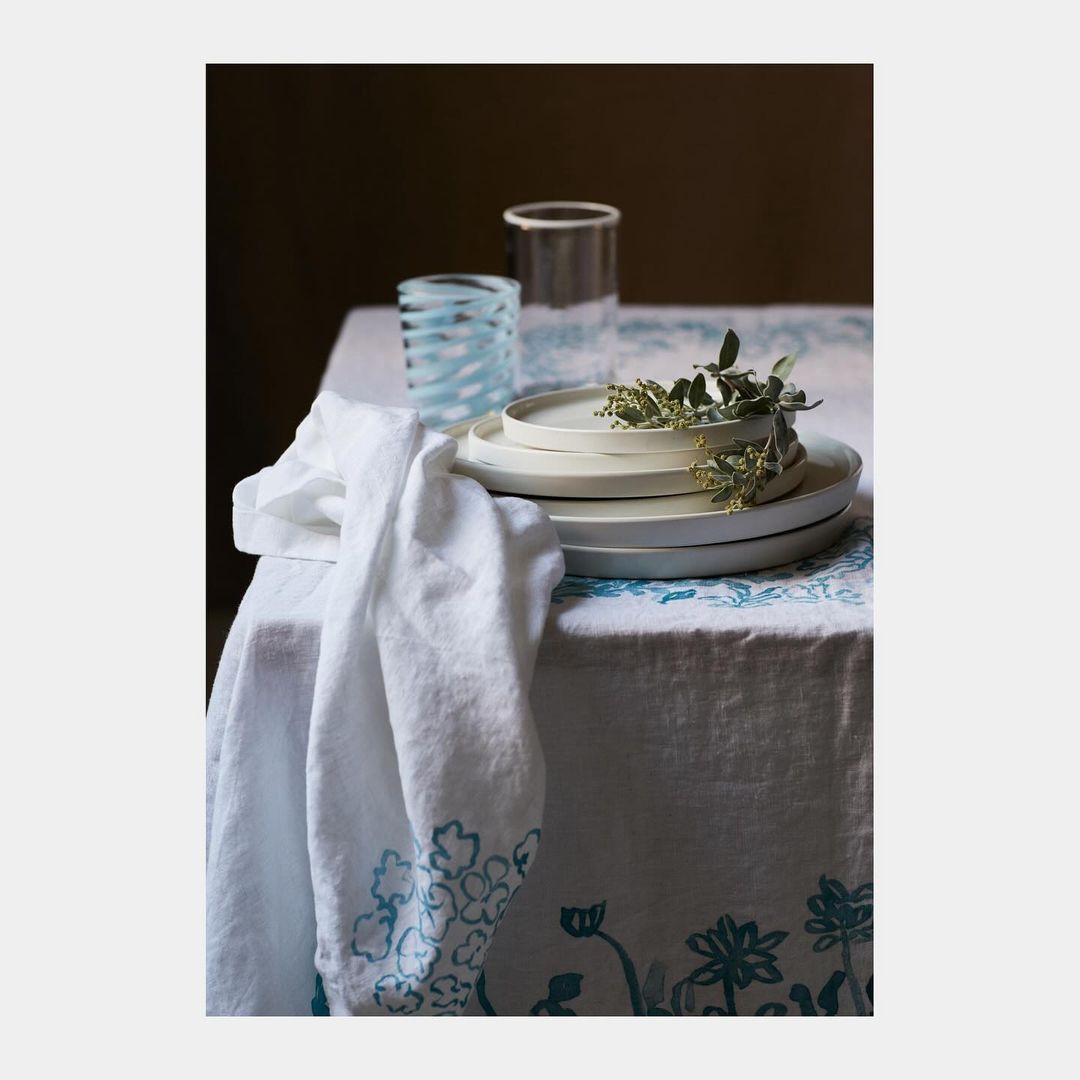 Still life shot of a linen-covered table with stacked ceramic plates, glassware, and a cloth, showcasing textures and muted tones.