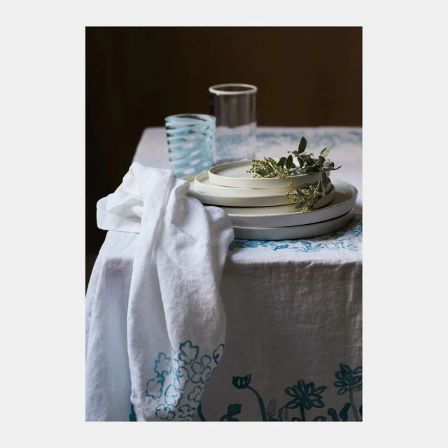Still life shot of a linen-covered table with stacked ceramic plates, glassware, and a cloth, showcasing textures and muted tones.