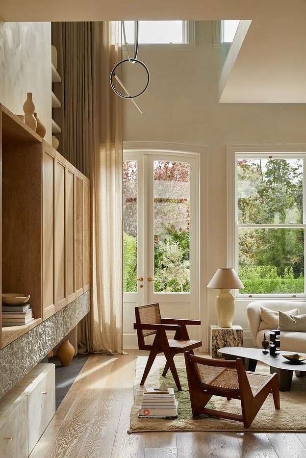 Bright interior shot of a living room with two wooden chairs, large windows overlooking a garden, and a built-in wooden cabinet.