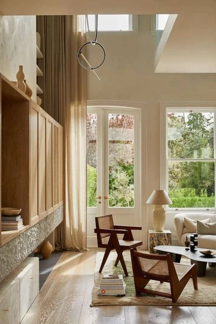 Bright interior shot of a living room with two wooden chairs, large windows overlooking a garden, and a built-in wooden cabinet.