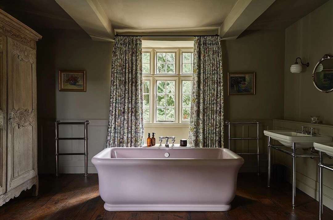 Interior view of a bathroom featuring a pink bathtub, floral curtains, and wooden floors. The space is naturally lit by a window.
