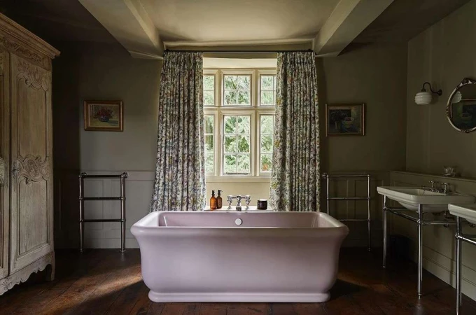 Interior view of a bathroom featuring a pink bathtub, floral curtains, and wooden floors. The space is naturally lit by a window.