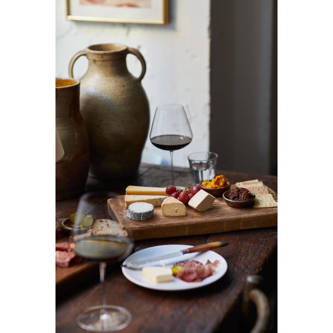 Still life shot of a wooden cutting board with various cheeses, grapes, and small bowls of condiments, accompanied by wine and water.