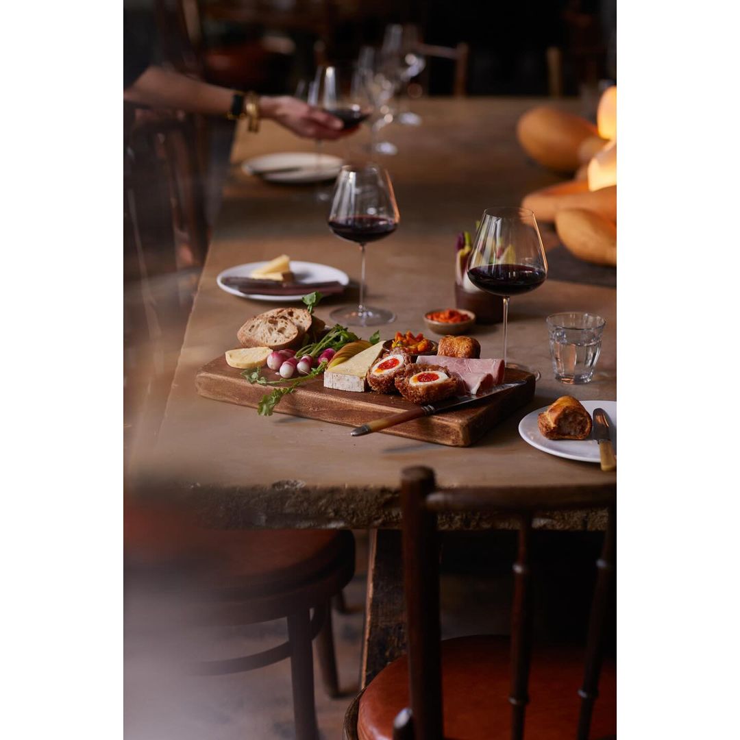 Wine glasses and a charcuterie board on a wooden table, showcasing cheeses, meats, and vegetables in a warm, inviting setting.