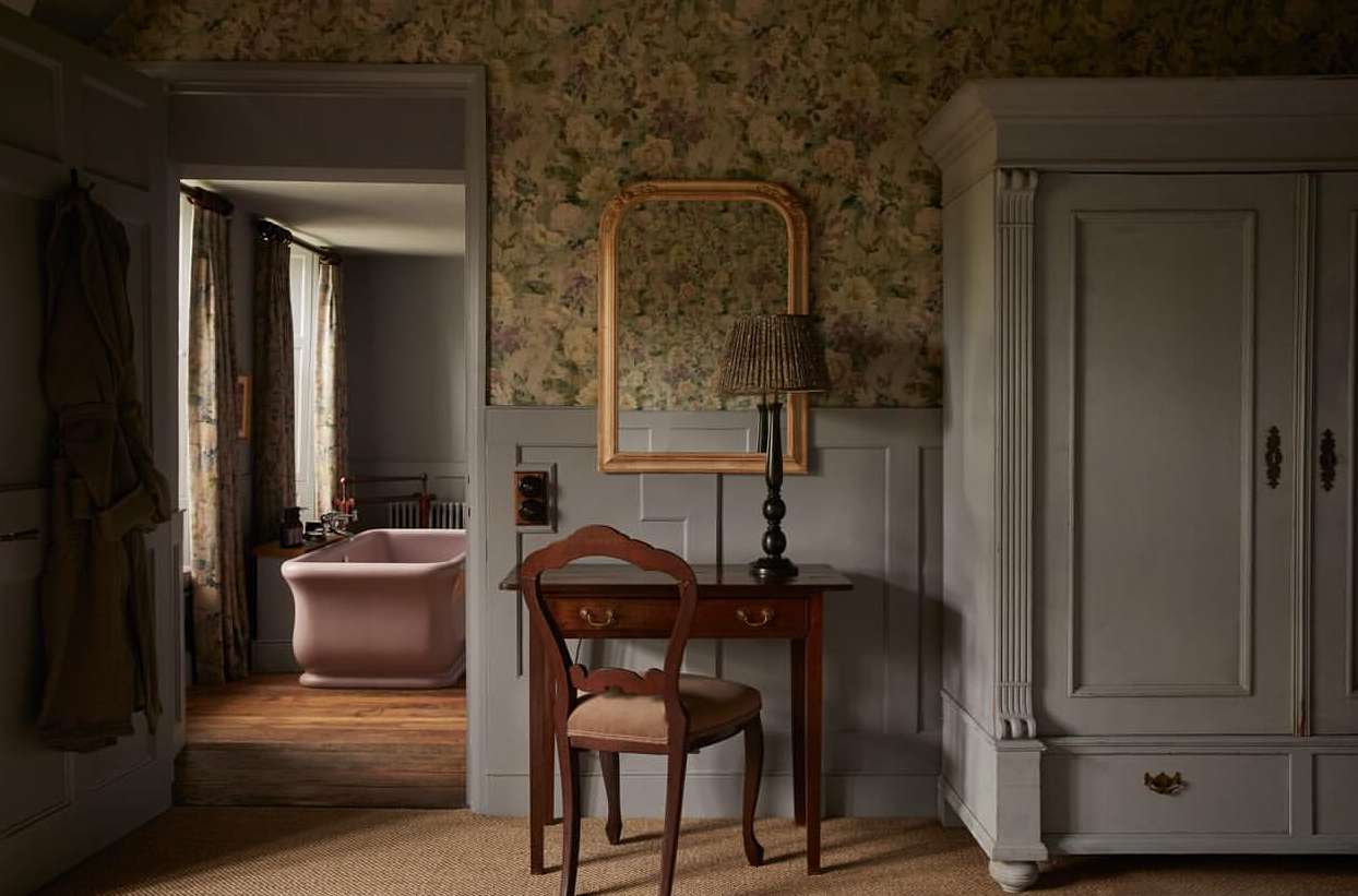 Bedroom interior featuring a vintage-style desk and chair, floral wallpaper, and a view into an adjacent bathroom with a pink bathtub.