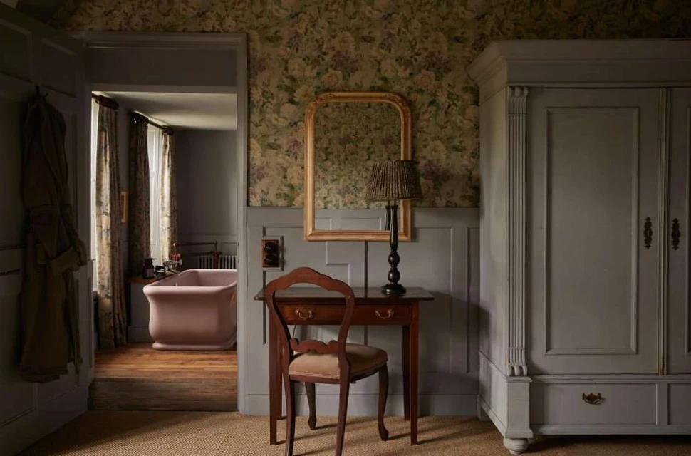 Bedroom interior featuring a vintage-style desk and chair, floral wallpaper, and a view into an adjacent bathroom with a pink bathtub.