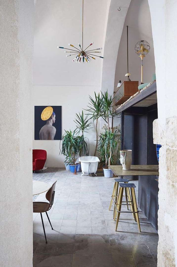 Interior of a room with a modern chandelier, plants, white bathtub, portrait on the wall, red chair, and several stools.