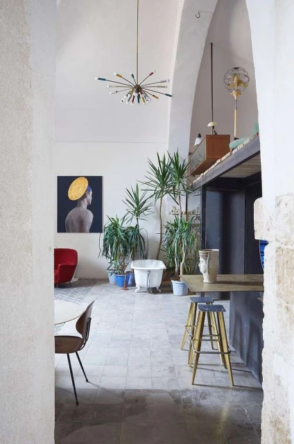 Interior of a room with a modern chandelier, plants, white bathtub, portrait on the wall, red chair, and several stools.