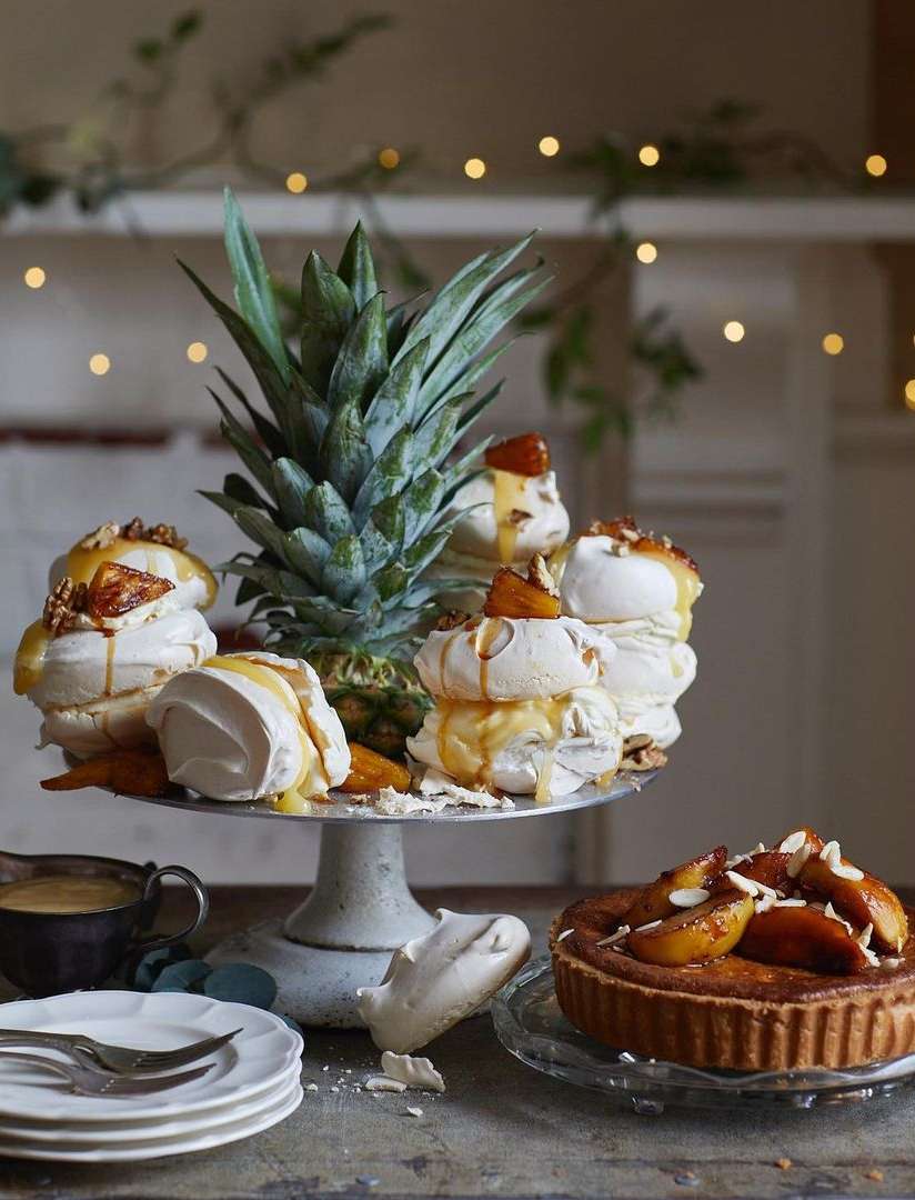 Close-up of meringue desserts, pineapple, and a tart arranged on stands and a table with a sauce boat and plates, warm lighting.