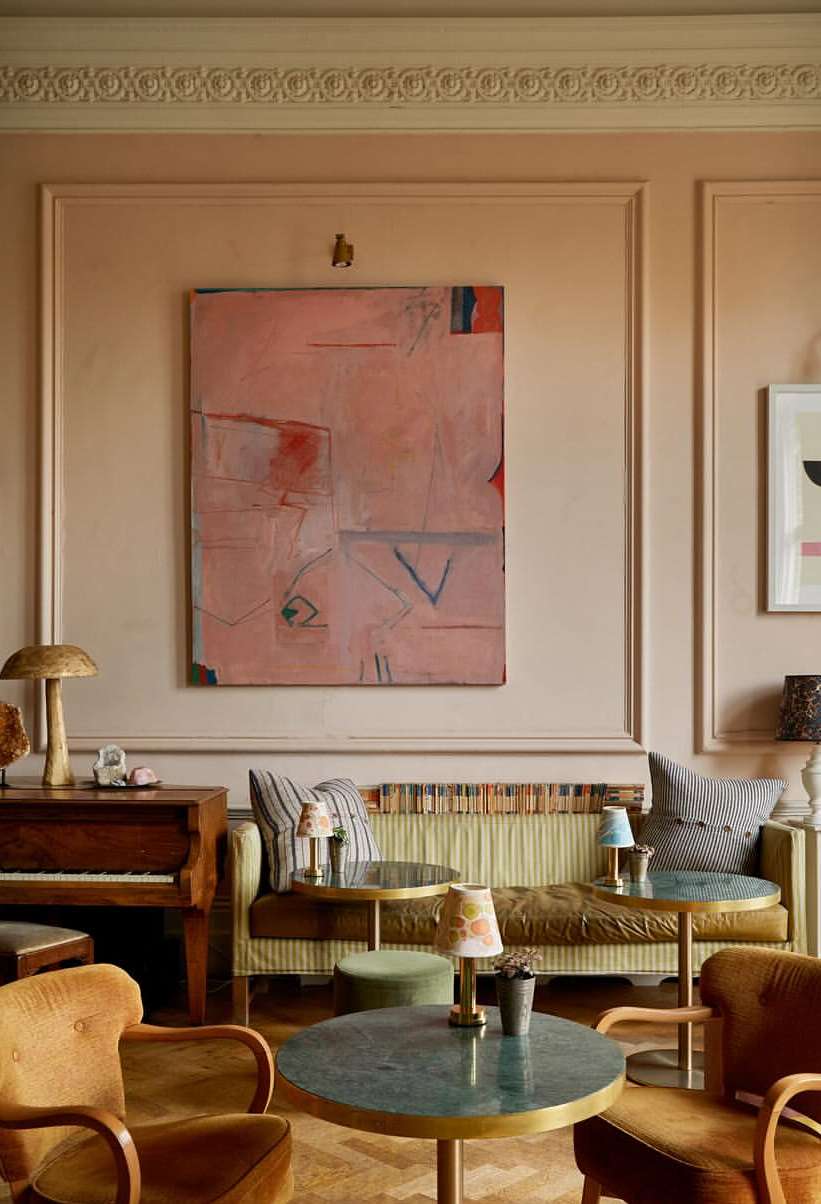 Interior view of a lounge area featuring a green couch, round tables, wood parquet flooring, an abstract painting, and two mustard-yellow armchairs.