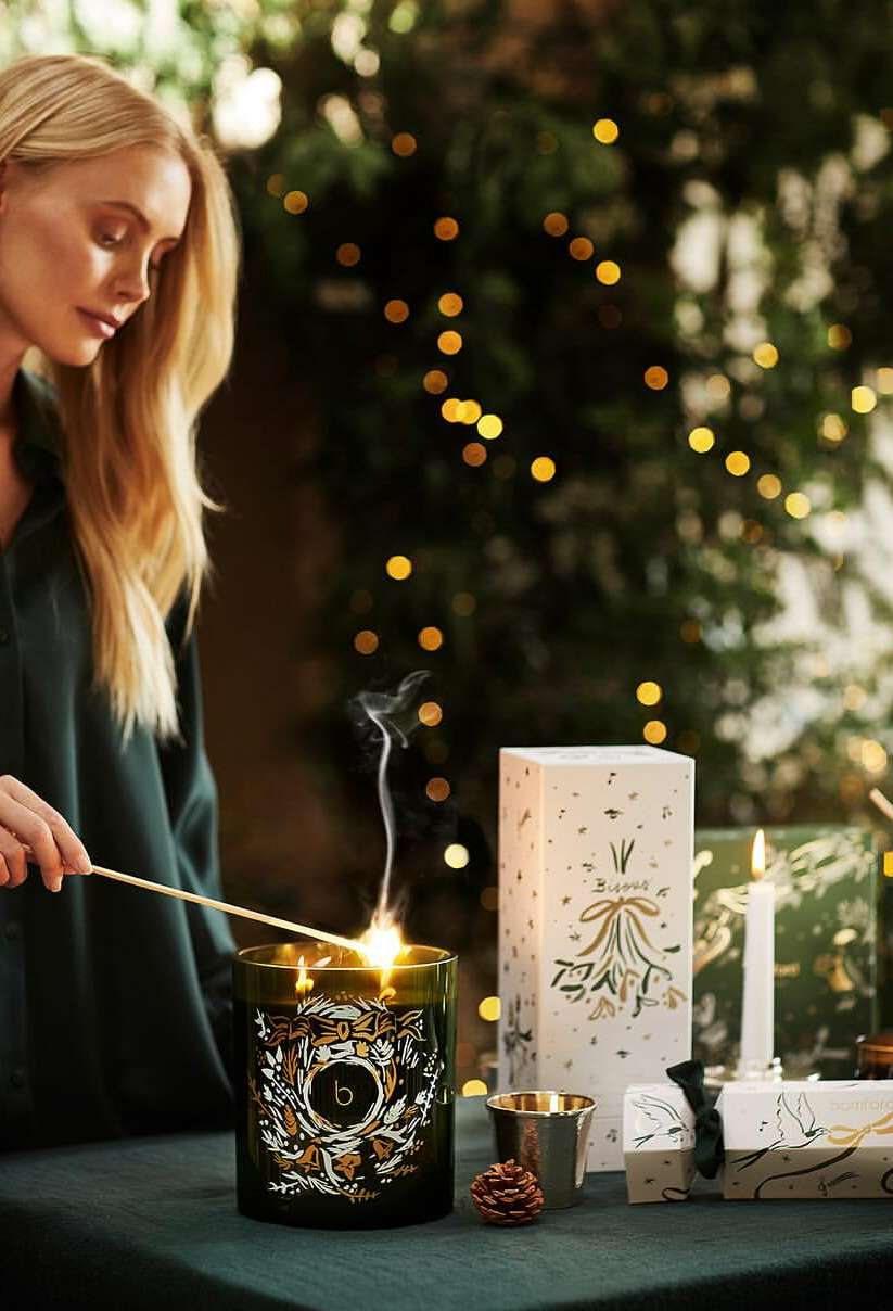 Blond woman lights candle with a match in an indoor setting. Gift boxes, holiday decor, and garland with lights are visible.