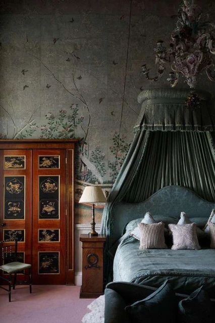 Elaborate canopy bed in an ornate bedroom featuring detailed wallpaper, decorative chandelier, and antique wooden closet with painted panels.