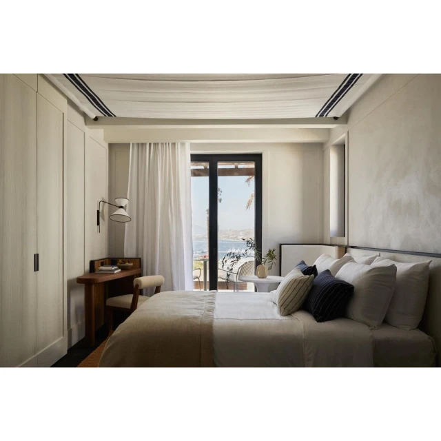 Tranquil bedroom with a made-up bed, wood desk, and ocean view through open balcony doors. Neutral color scheme, natural light.