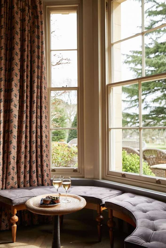 Cozy window seat with two wine glasses and snacks on a round table, set in a bright, inviting room with an outdoor garden view.