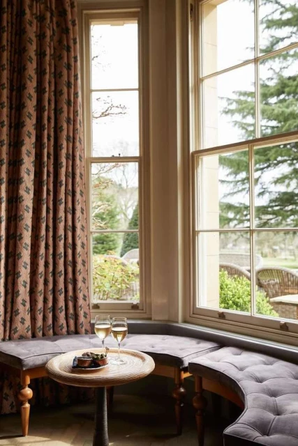 Cozy window seat with two wine glasses and snacks on a round table, set in a bright, inviting room with an outdoor garden view.