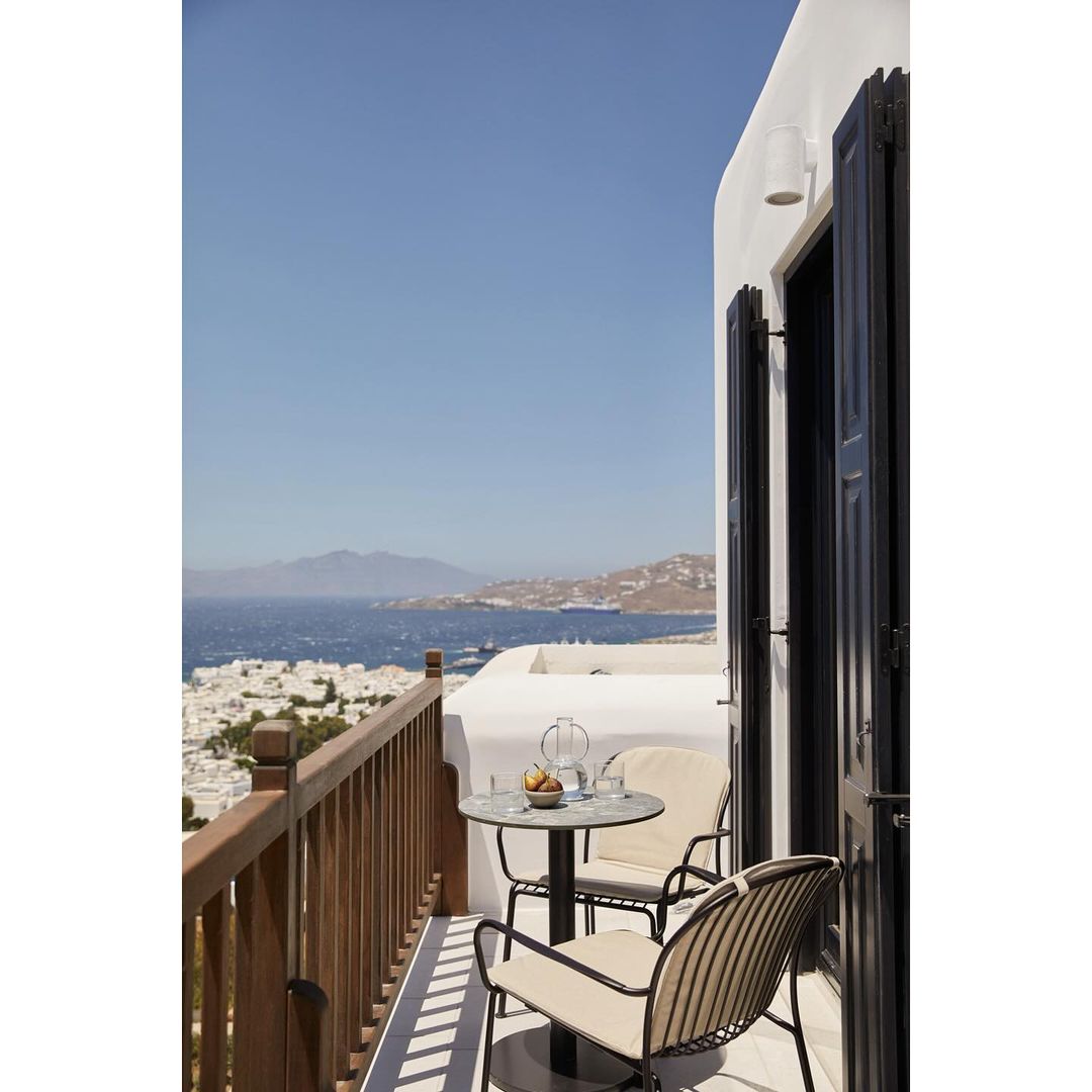 Outdoor balcony with seating featuring a table and chairs on a sunny day, overlooking the sea and distant hills, creating a relaxing atmosphere.