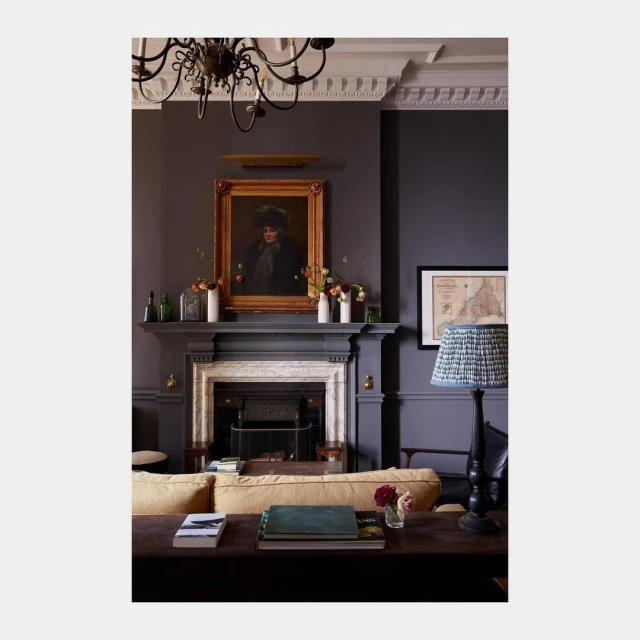 Interior view of a living room with dark grey walls, a fireplace, a painting above the mantel, books on a table, and a floor lamp.