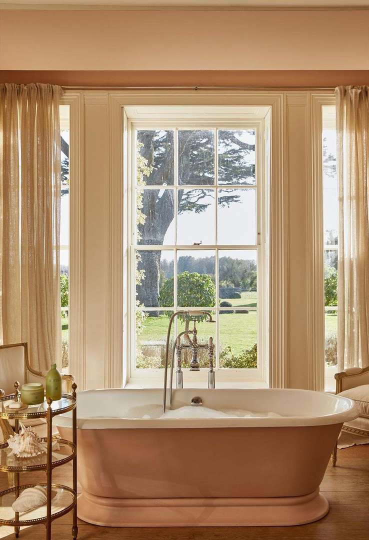 Elegant bathroom featuring a freestanding peach-colored bathtub filled with bubbles, set against a bright window view of lush greenery.