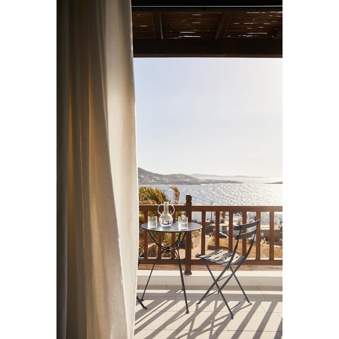 Outdoor balcony with table and chair overlooking ocean. Clear glass pitcher and glasses on table. Day time. Balcony has wooden railing.
