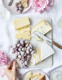 Overhead food photography of a marble cheese board with assorted cheeses, grapes, and pink flowers on white linen.