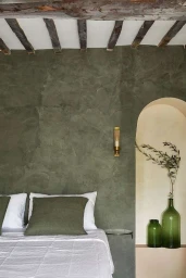 Interior bedroom view showing an olive green textured wall, white bed linens, and green glass bottles in an arched niche by Helen Cathcart.