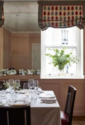 Interior shot of an elegantly set restaurant dining table with white linens and glassware in front of a window.